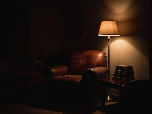 Deep-shadow photograph of a comfortable armchair beside a warm floor lamp and a stack of books in a quiet evening living room