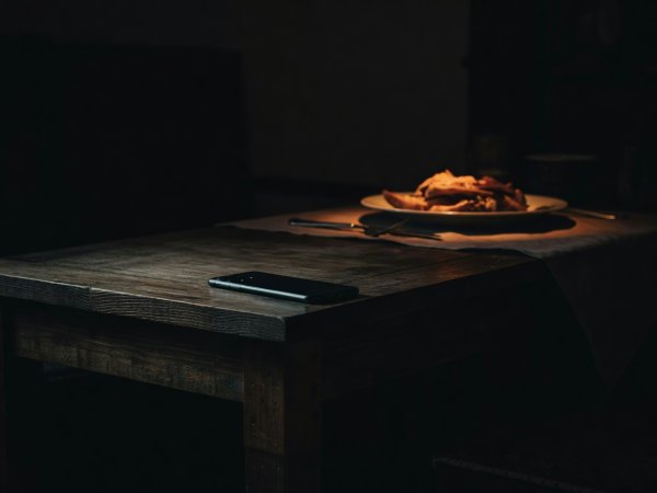 High-contrast photograph of a smartphone resting face down on a wooden side table away from a warm dinner plate in deep evening shadows
