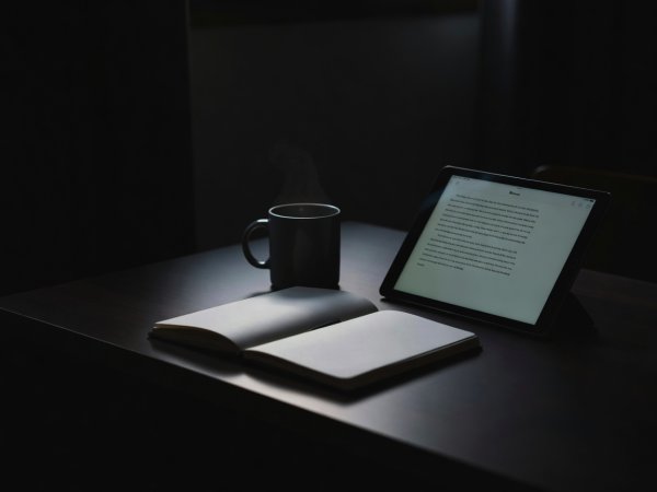 High-contrast photograph of an open notebook and ceramic mug beside a tablet showing a clean text reader in deep morning shadow